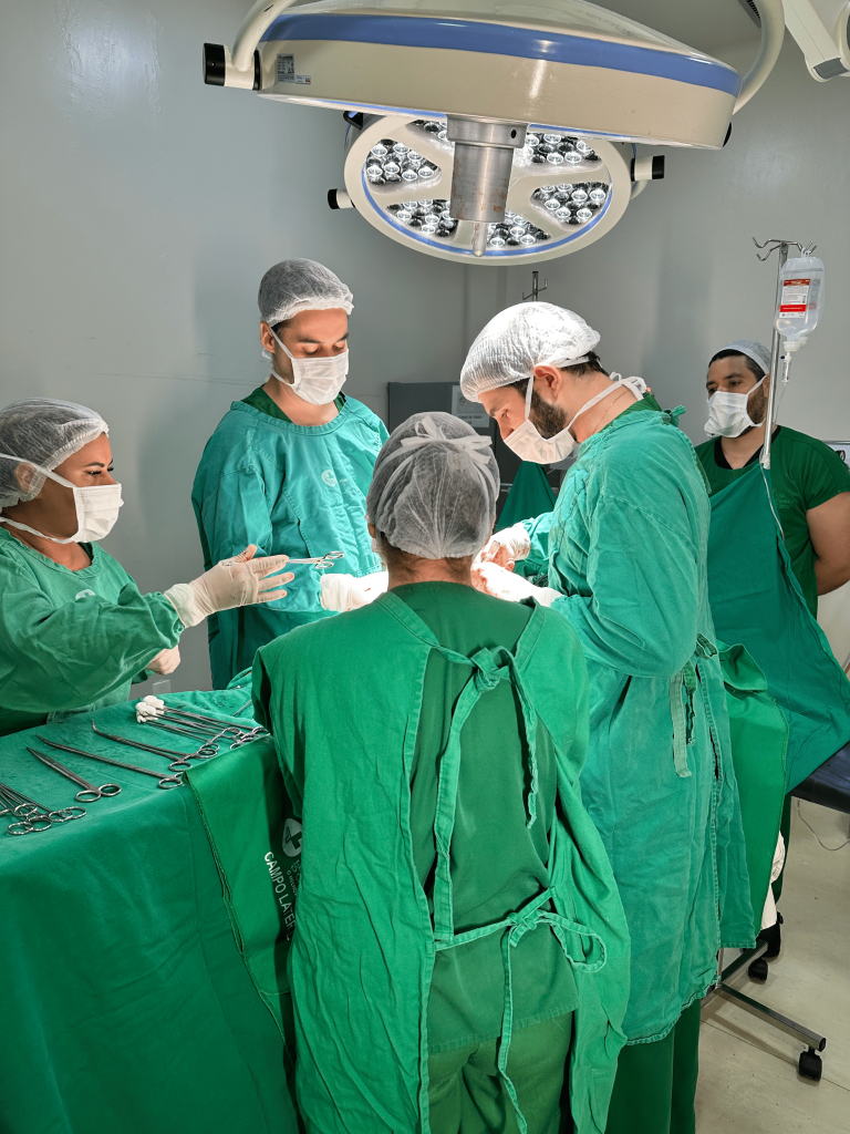 Imagem de cirurgia com dois cirurgiões urológicos um de frente para o outro, com o paciente deitado e inconsciente na maca, ao centro. Há também duas enfermeiras auxiliando. Acima do paciente há uma luminária redonda de fácil rotação. Ao fundo da foto, o médico anestesista assiste a operação concentrado, próximo ao soro do paciente.