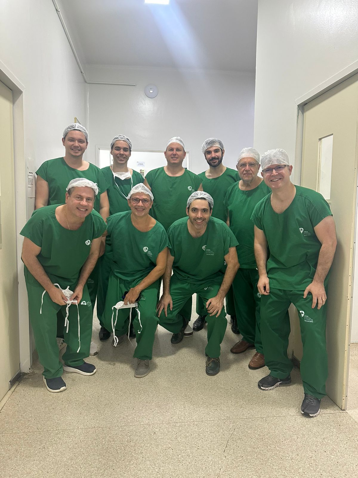 No corredor do centro cirúrgico, estão os nove cirurgiões urologistas de toucas no cabelo, sem máscara, sorrindo para a foto. Todos vestem os "pijamas cirúrgicos" da Liga Contra o Câncer. 
