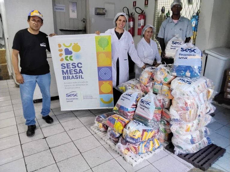 A imagem mostra um conjunto de profissionais de nutrição, com jaleco e touca, recebendo várias cestas básicas com o nome Sesc Mesa Brasil. Ao todo são 610 quilogramas de alimentos, como feijão, arroz, cuscuz, entre outros.