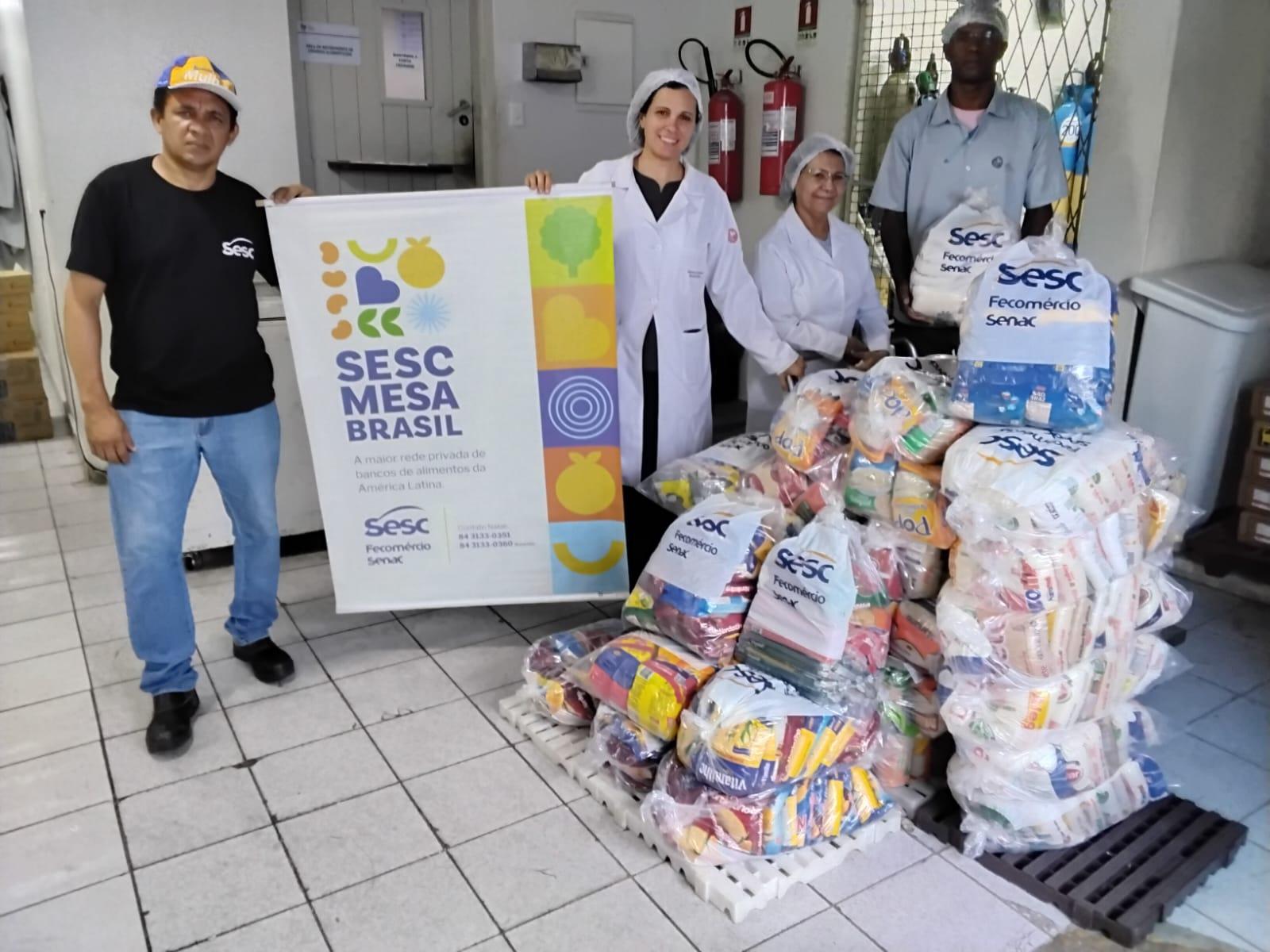 A imagem mostra um conjunto de profissionais de nutrição, com jaleco e touca, recebendo várias cestas básicas com o nome Sesc Mesa Brasil. Ao todo são 610 quilogramas de alimentos, como feijão, arroz, cuscuz, entre outros.